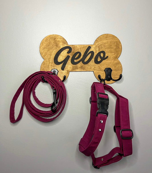 A personalized dog accessory hanger with a golden bone-shaped sign that reads 'Gebo', mounted on a wall with a red leash and a black harness.