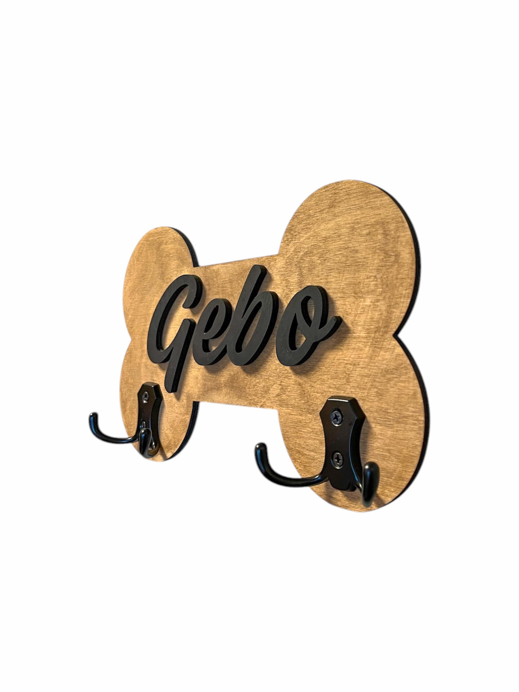 A personalized dog accessory hanger with a golden bone-shaped sign that reads 'Gebo', mounted on a wall with a red leash and a black harness.